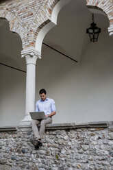 Geschäftsmann sitzt auf einer Steinmauer und benutzt einen Laptop - MAUF000547