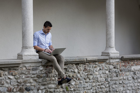 Geschäftsmann sitzt auf einer Steinmauer und benutzt einen Laptop, lizenzfreies Stockfoto