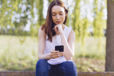 Young woman with phone sitting on a bench at the park - GIOF000933