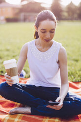 Student im Park, der einen Kaffee zum Mitnehmen trinkt und auf sein Smartphone schaut - GIOF000955