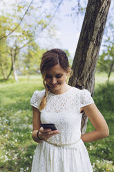 Lächelnde Frau lehnt an einem Baumstamm und schaut auf ihr Smartphone - GIOF000978