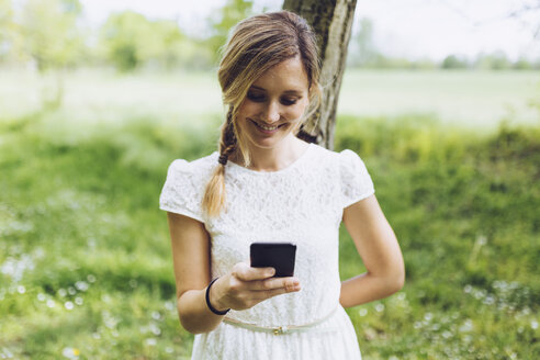 Lächelnde Frau lehnt an einem Baumstamm und schaut auf ihr Smartphone - GIOF000979