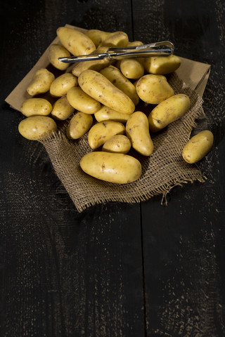 Rohe Cherie-Kartoffeln und Schälmaschine auf braunem Papier, Jute und dunklem Holz, lizenzfreies Stockfoto