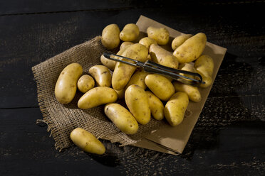 Raw Cherie potatoes and peeler on brown paper, jute and dark wood - MAEF011667