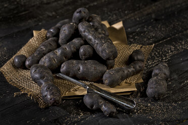 Blue violet potatoes and peeler on brown paper, jute and dark wood - MAEF011668