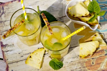 Two glasses with ice, cocos sirup and ananas juice, garnished with pieces of ananas and lemon balm - YFF000542