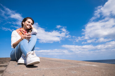 Lächelndes Teenager-Mädchen sitzt an der Wand an der Küste und hört Musik - SIPF000455