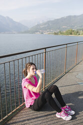 Italy, Lecco, sportive young woman drinking water after running at lakeshore - MRAF000028