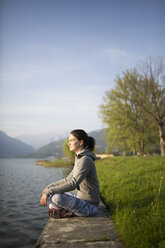 Italy, Lecco, relaxed young woman sitting at the lakeshore - MRAF000041