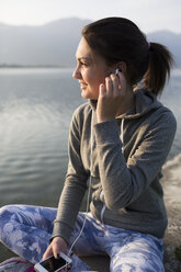 Italy, Lecco, smiling young woman sitting at the lakeshore listening to music - MRAF000046