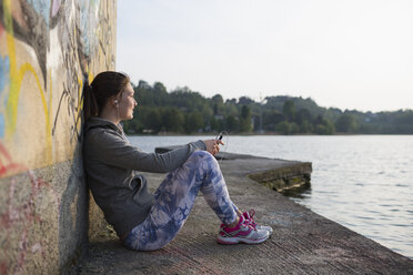 Italien, Lecco, entspannte junge Frau sitzt am Seeufer und hört Musik - MRAF000048