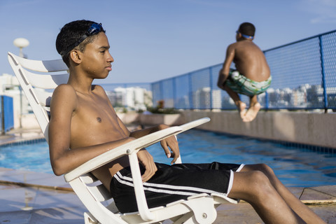 Teenager sitzt auf einem Stuhl vor dem Schwimmbad, während sein Freund einen Kanonenkugelsprung macht, lizenzfreies Stockfoto