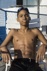 Teenage boy sitting on chair outside swimming pool - MAUF000572
