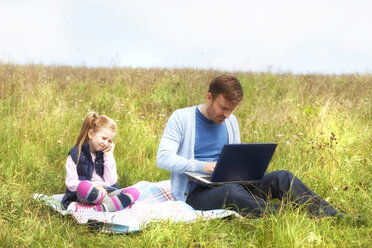 Father and daughter sitting on meadow, smartphone and laptop - MAEF011727