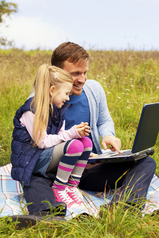Vater und Tochter sitzen auf einer Wiese, der Vater arbeitet am Laptop, lizenzfreies Stockfoto