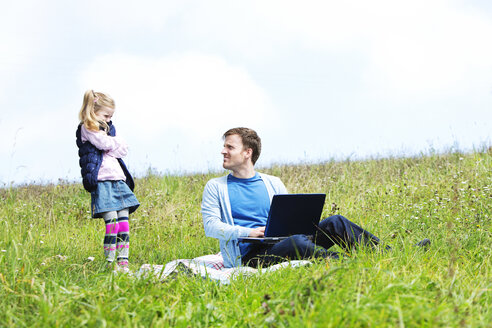 Vater und Tochter sitzen auf einer Wiese, Laptop, Vater schaut zur Tochter - MAEF011732
