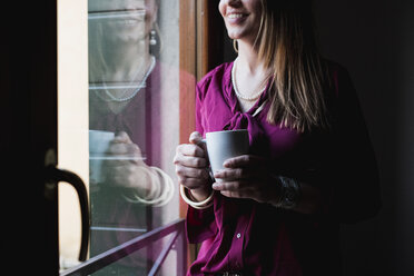 Young woman standing by window, drinking coffee - MAUF000583