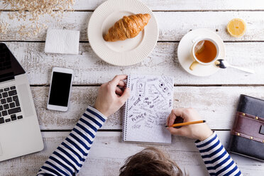 Man working at table with croissant and tea, taking notes - HAPF000358