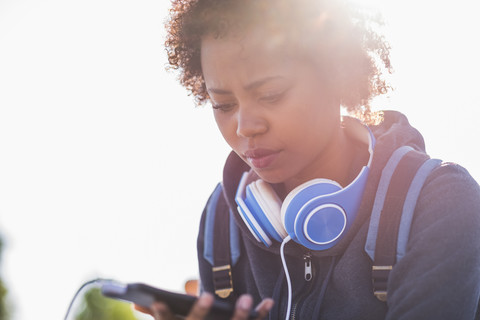 Junge Frau mit Kopfhörern, die im Freien auf ihr Mobiltelefon schaut, lizenzfreies Stockfoto