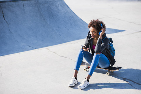 Junge Frau sitzt auf einem Skateboard und hört Musik, lizenzfreies Stockfoto