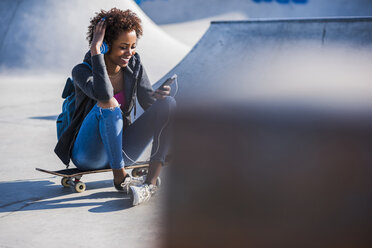 Lächelnde junge Frau sitzt auf einem Skateboard und hört Musik - UUF007248