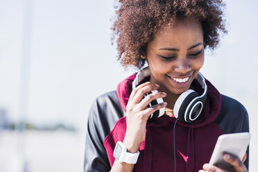 Lächelnde junge Frau mit Kopfhörern, die im Freien auf ihr Mobiltelefon schaut - UUF007303