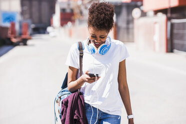 Smiling young woman wearing headphones looking at cell phone outdoors - UUF007307