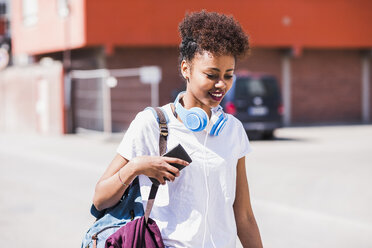 Lächelnde junge Frau mit Kopfhörern und Handy in der Hand im Freien - UUF007308