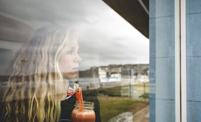 Porträt einer lockigen blonden Frau, die durch das Fenster eines Cafés schaut und einen Saft in den Händen hält - DAPF000104