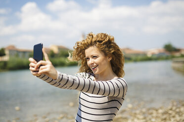 Italien, Verona, Porträt einer lächelnden Frau, die am Flussufer ein Selfie mit ihrem Smartphone macht - GIOF001040