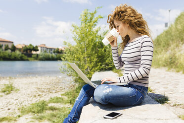 Woman drinking coffee to go while using laptop at riverside - GIOF001041