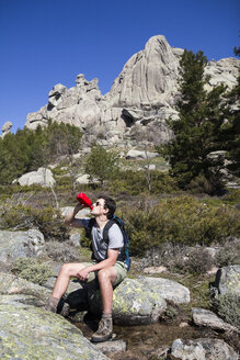 Spanien, Wanderer mit Sonnenbrille und Rucksack, der aus einer Flasche trinkt, die auf einem Felsen in La Pedriza steht - ABZF000544