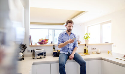 Mann sitzt in der Küche und benutzt ein Smartphone - HAPF000396