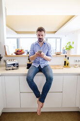 Mann sitzt in der Küche und benutzt ein Smartphone - HAPF000397