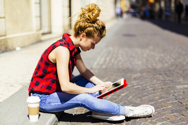 Italy, Verona, woman sitting on curb using digital tablet - GIOF001065