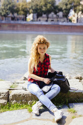 Italy, Verona, smiling woman sitting at the riverside - GIOF001090