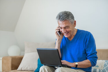 Smiling senior man sitting on couch using laptop and cell phone - DIGF000496