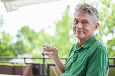 Portrait of smiling senior man sitting on balcony holding glass of water - DIGF000518