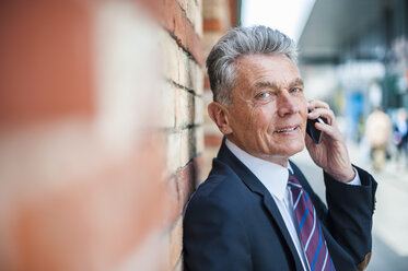 Älterer Geschäftsmann lehnt an einer Backsteinmauer und spricht mit einem Handy - DIGF000539