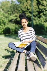 Porträt einer jungen Frau, die mit einem Buch auf einer Parkbank sitzt - GIOF001096