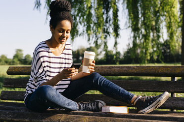 Junge Frau sitzt auf einer Parkbank mit einem Buch und einem Kaffee und schaut auf ihr Smartphone - GIOF001098
