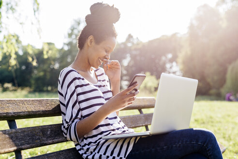 Laughing young woman sitting on park bench with smartphone and laptop - GIOF001105