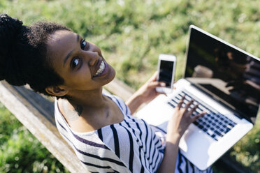 Porträt einer lächelnden Frau, die auf einer Parkbank sitzt und einen Laptop und ein Smartphone benutzt - GIOF001107