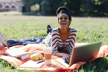 Lachende Frau mit Laptop und Kaffee zum Mitnehmen auf einer Decke auf einer Wiese liegend - GIOF001108