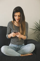 Junge Frau sitzt im Schneidersitz auf der Couch und benutzt ein Smartphone - EBSF001417