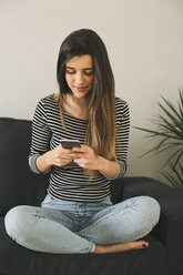 Young woman sitting cross-legged on couch, using smart phone - EBSF001417