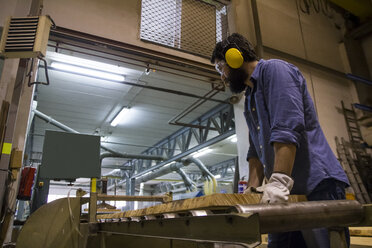 Handwerker mit Gehörschutz, Handschuhen und Schutzbrille bei der Arbeit mit einer Industriekreissäge in einer Fabrik - ABZF000553