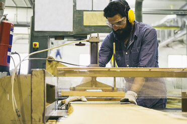 Handwerker mit Gehörschutz, Handschuhen und Schutzbrille bei der Arbeit mit einer Industriekreissäge in einer Fabrik - ABZF000554