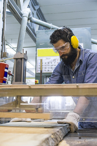 Handwerker mit Gehörschutz, Handschuhen und Schutzbrille bei der Arbeit mit einer Industriekreissäge in einer Fabrik, lizenzfreies Stockfoto