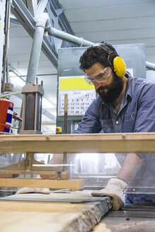 Craftsman with hearing protection, gloves and safety glasses using an industrial circular saw in a factory - ABZF000555